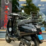 Zwarte scooter op middenbok voor een winkelraam met Kymco-banner en reflecties van bomen.