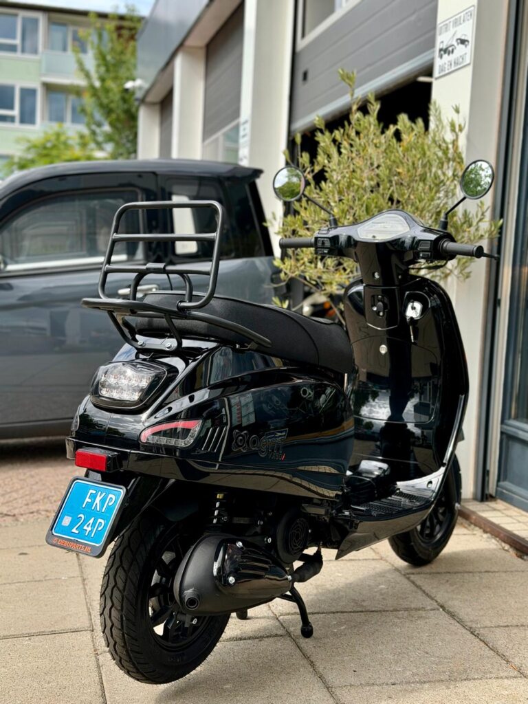 Zwarte scooter met bagagedrager geparkeerd voor werkplaats met open roldeuren.