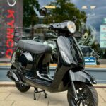 Zwarte retro scooter op standaard voor een winkelruit met KYMCO-vlag op de achtergrond.