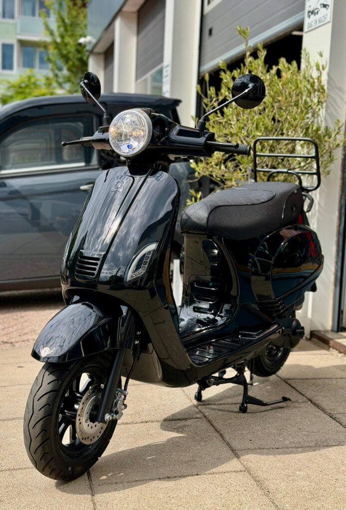 Zwarte scooter met ronde koplamp en bagagerek, geparkeerd op stoep bij garage.