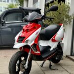 Rood-witte scooter op standaard geparkeerd op stoep voor een werkplaats met grijze bestelwagen.