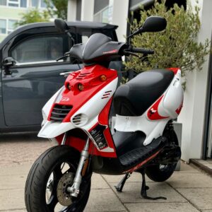 Rood-witte scooter op standaard geparkeerd op stoep voor een werkplaats met grijze bestelwagen.