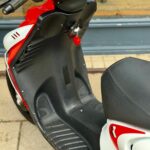 Close-up van een rood-witte scooter met voetplaat, zadel en sleutel in het contact.