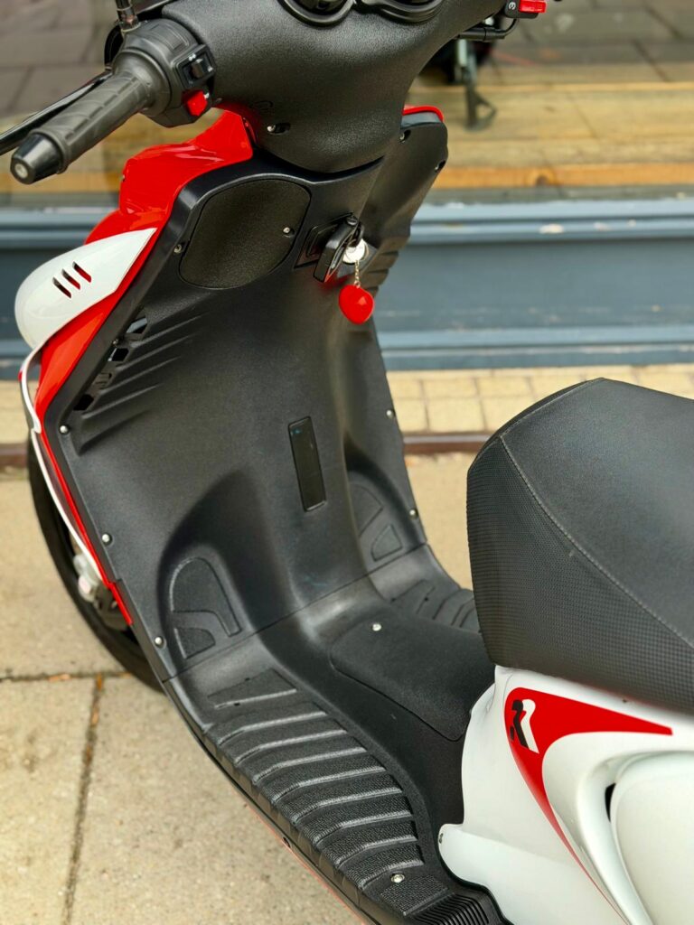 Close-up van een rood-witte scooter met voetplaat, zadel en sleutel in het contact.
