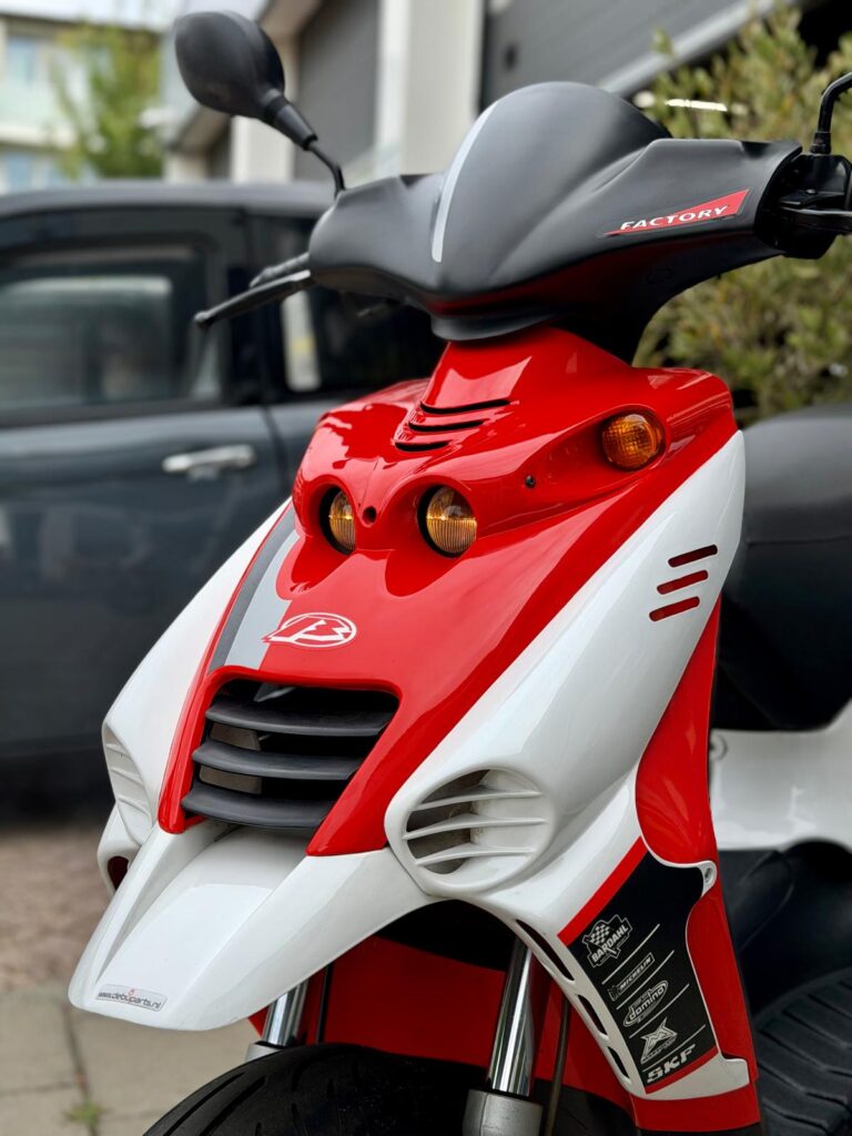 Close-up van een rode-witte sportieve scooter met koplampen en stickers, geparkeerd buiten.