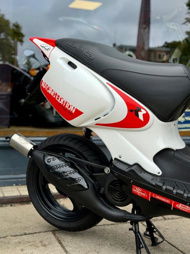 Achterzijde van een witte en rode Beta Racing Edition scooter met zwarte uitlaat, geparkeerd voor een winkelruit.