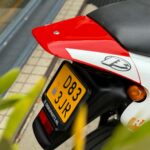 Achterzijde van een sportieve motor met rood spatbord, zwart zadel en gele NL-kentekenplaat bij een stoep.