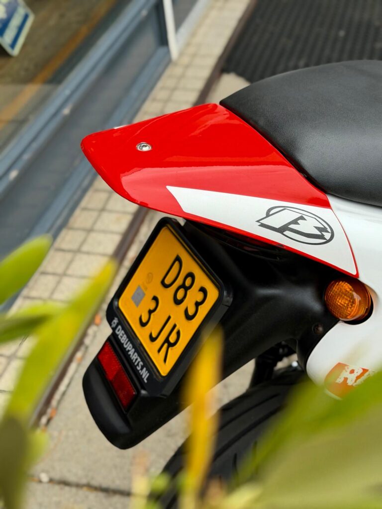 Achterzijde van een sportieve motor met rood spatbord, zwart zadel en gele NL-kentekenplaat bij een stoep.