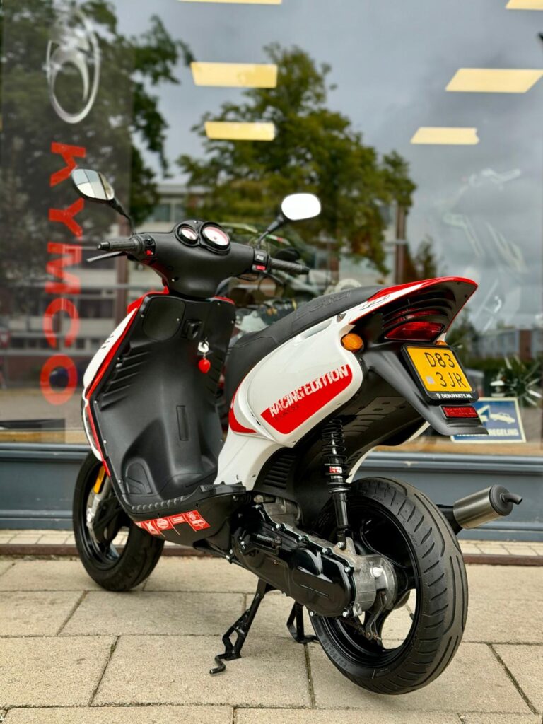 Wit-rode Kymco-scooter met Racing Edition, geparkeerd voor een winkelruit op de stoep.
