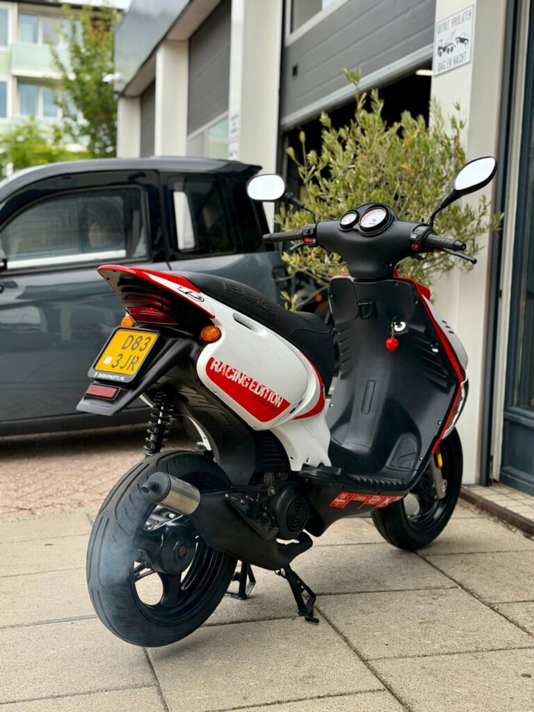 Wit-rode Racing Edition scooter op zijstandaard voor een garage, achteraanzicht met gele kentekenplaat.
