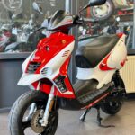 Rood-witte scooter op standaard in een showroom met andere scooters op de achtergrond.