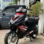 Zwarte Yamaha-scooter met rode accenten en witte velgen, geparkeerd op de stoep bij een garage.