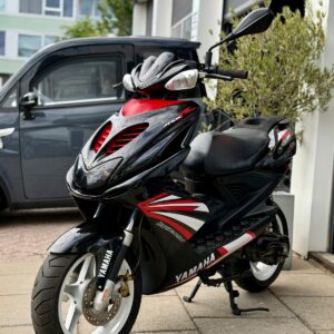 Zwarte Yamaha-scooter met rode accenten en witte velgen, geparkeerd op de stoep bij een garage.