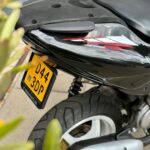 Close-up van de achterzijde van een zwarte scooter met gele kentekenplaat, dikke band en zilveren uitlaat.