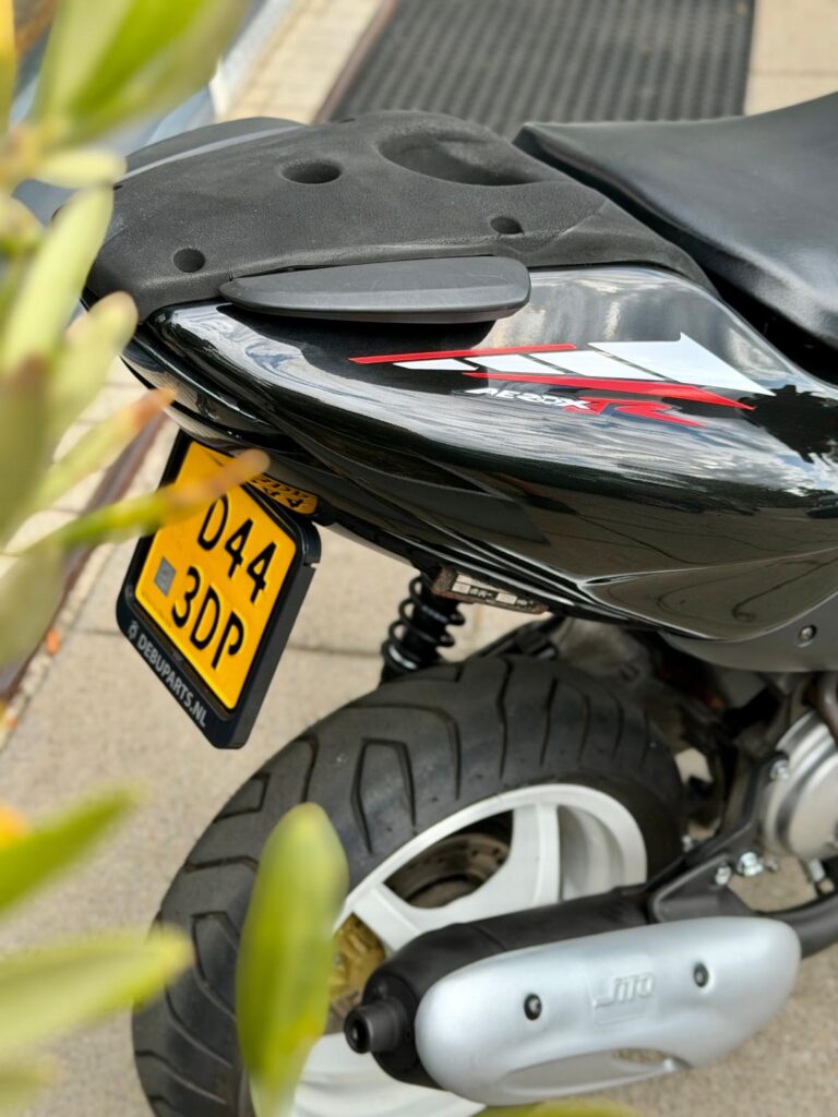 Close-up van de achterzijde van een zwarte scooter met gele kentekenplaat, dikke band en zilveren uitlaat.