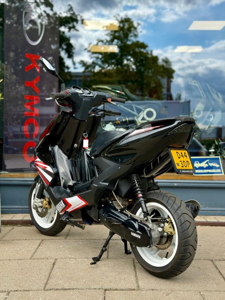 Zwarte Yamaha scooter met witte velgen, geparkeerd voor een winkel met Kymco-logo op het raam.