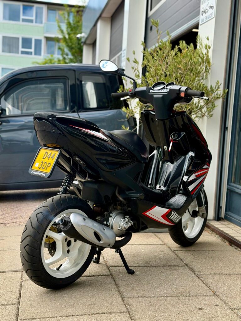 Zwarte Yamaha-scooter met witte velgen geparkeerd op stoep bij garagebox.