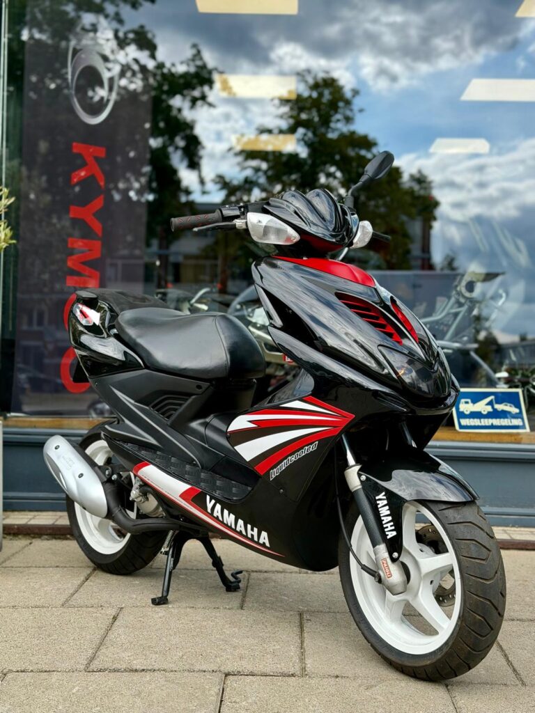 Zwarte en rode Yamaha scooter op de stoep, geparkeerd voor een winkelruit met reflecties.